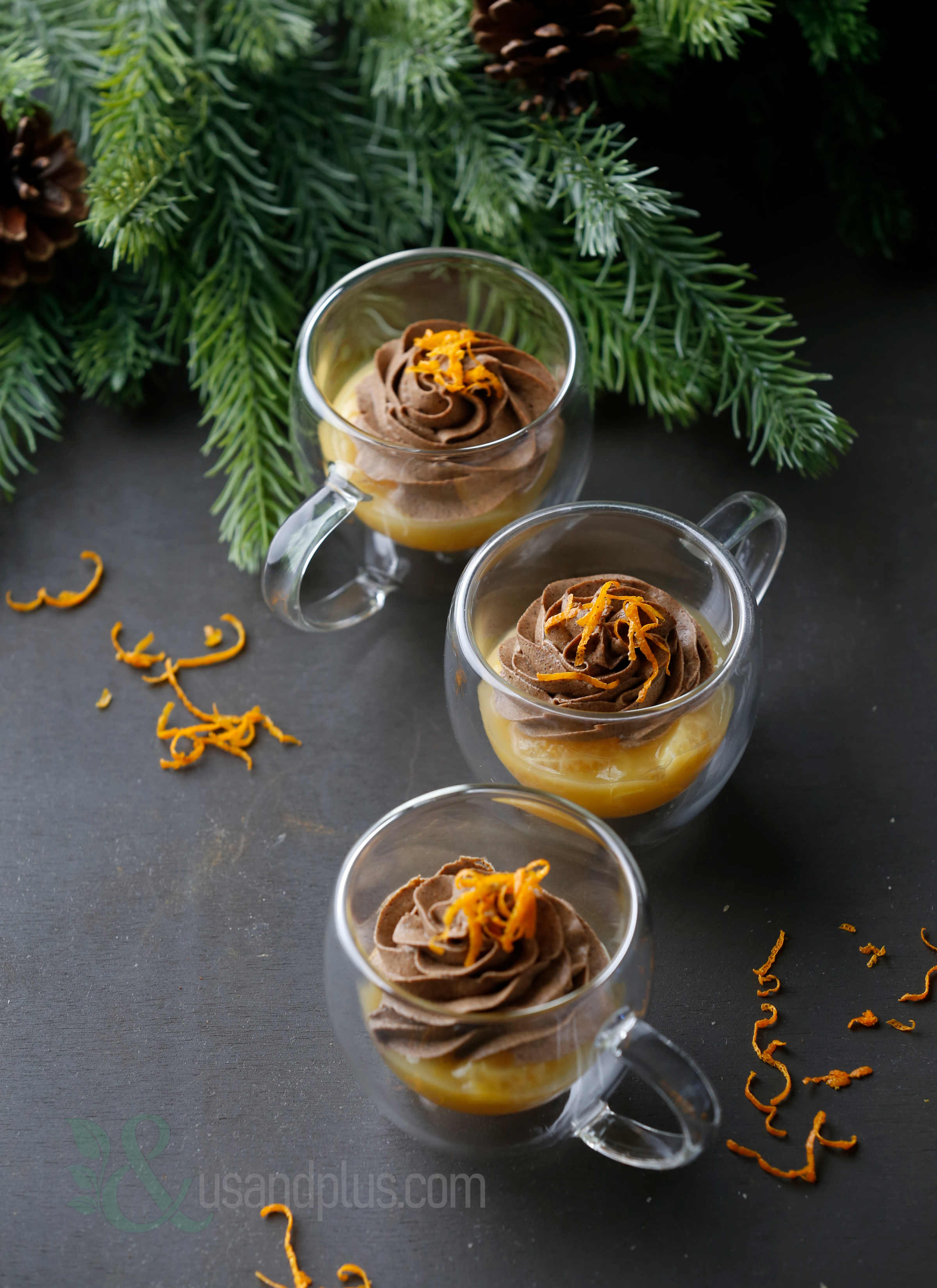 Three clear glass cups containing dessert with chocolate frosting and orange zest, surrounded by pine branches and orange peel on a dark surface.